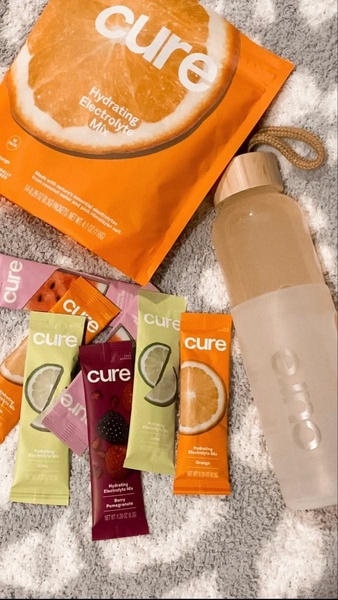 A high-quality product hero shot featuring assorted electrolyte mix packets and a branded reusable water bottle on a soft, textured background.